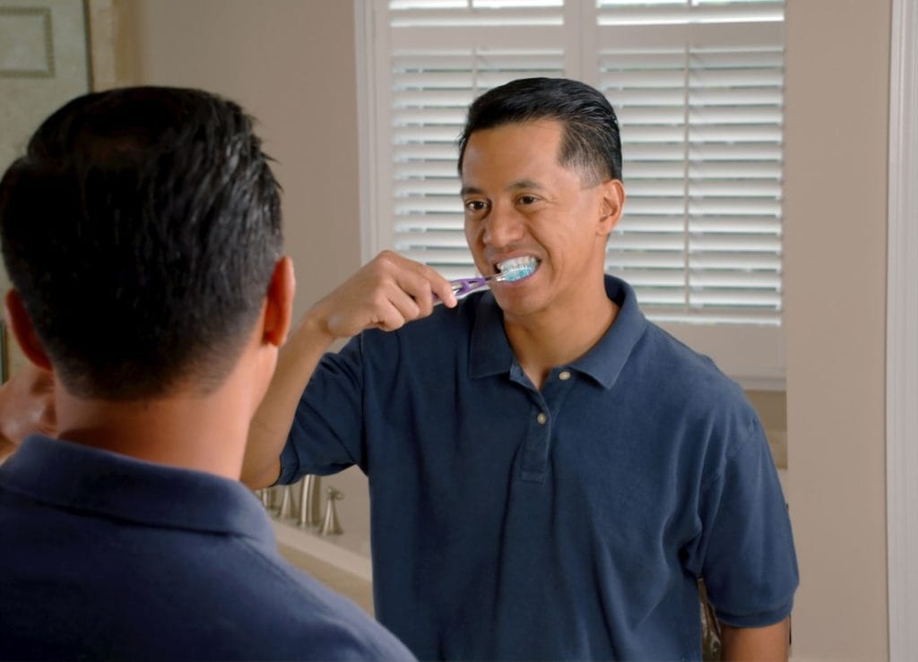 An Asian man brushing his teeth while looking in a mirror.