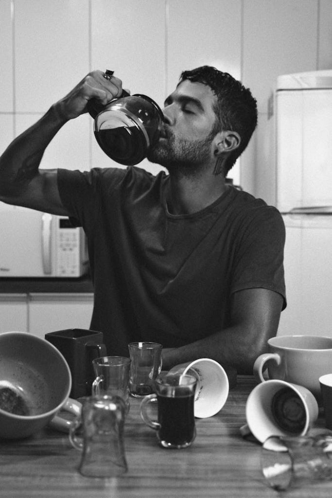 About Black and white photo of a man drinking coffee straight from a pitcher at the kitchen table.